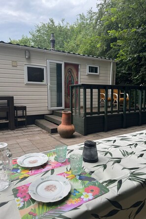 Ein Tisch draußen gedeckt mit Tellern, Gläsern und einer Vase. Im Hintergrund ist ein kleines Gebäude mit einer Tür und Fenstern zu sehen.