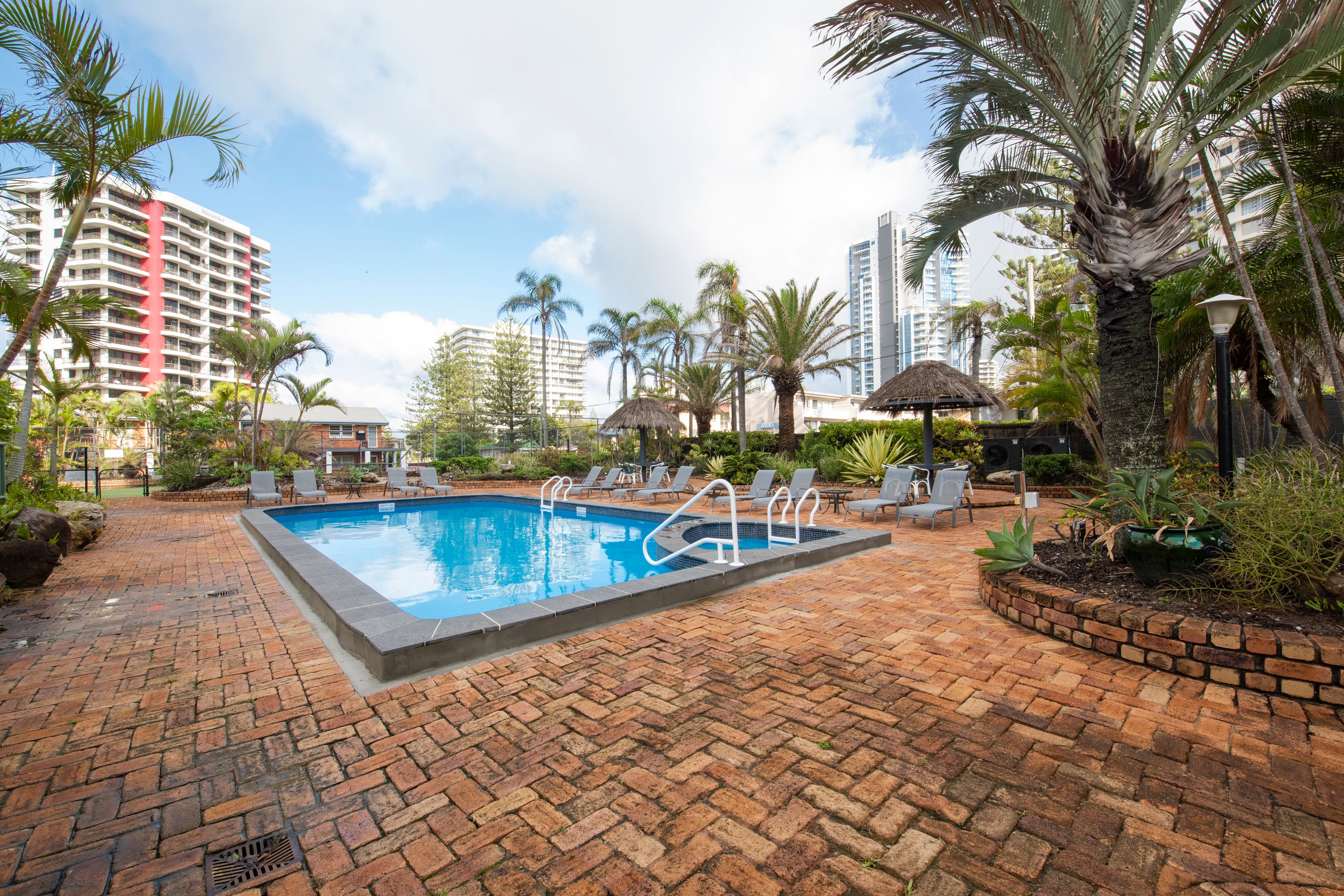 A swimming pool with lounge chairs and palm trees, surrounded by brick walkways and tall buildings.