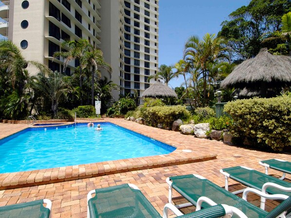 Una piscina con sillas de descanso, palmeras y un edificio alto al fondo.