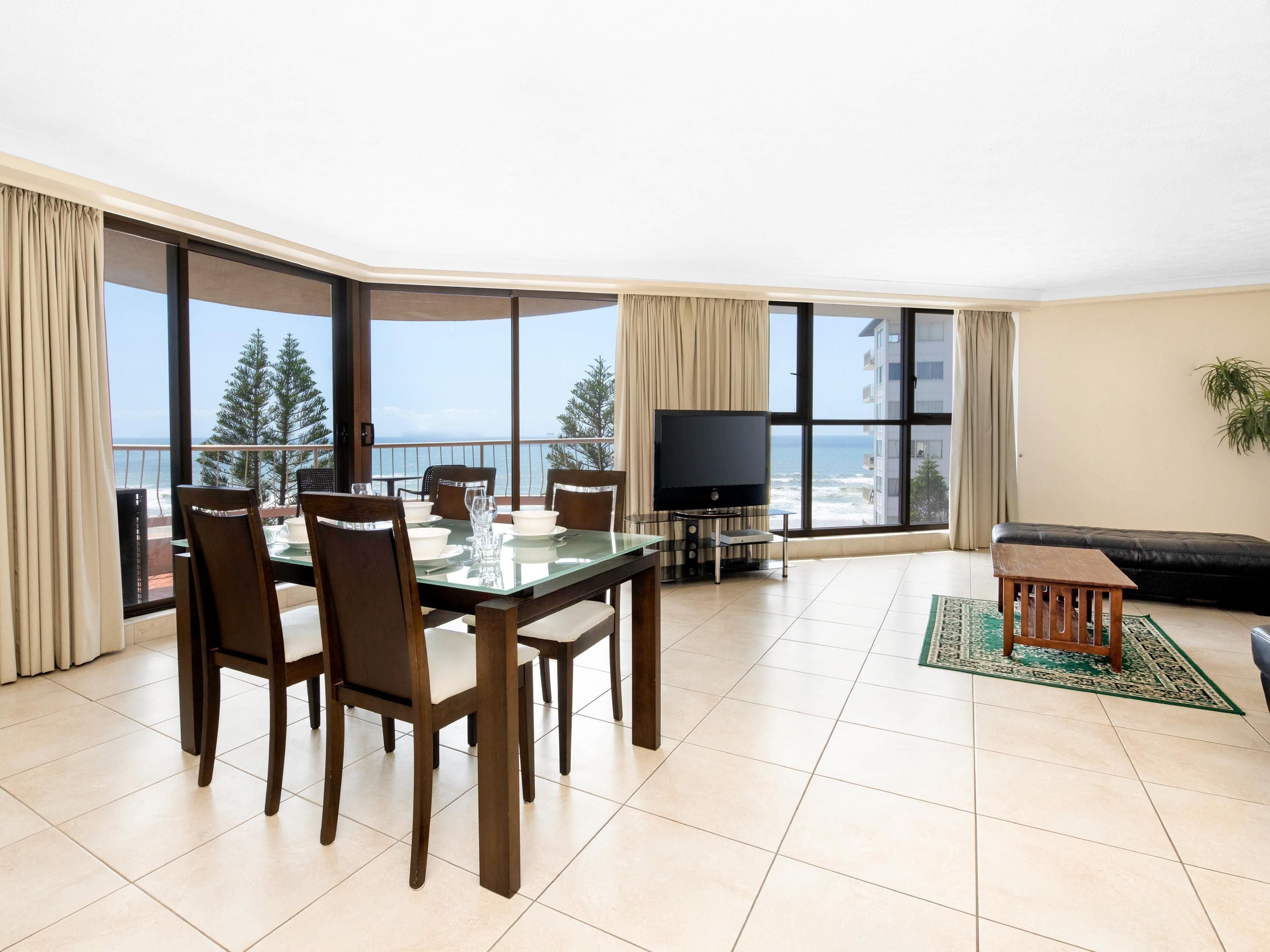 A modern living room with a dining area, large windows, a flat-screen TV, and a view of the ocean.