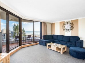 A modern living room with a large window, a blue sofa, a wooden coffee table, and a decorative wall clock. - Breakfree Acapulco Surfers Paradise (Gold Coast)