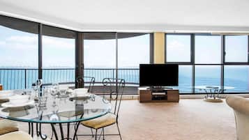 A modern living room with a glass table, chairs, a flat-screen TV, and a view of the ocean.