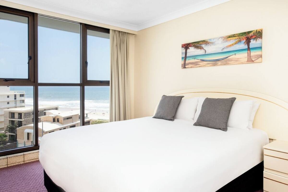 A hotel room with a large bed, a painting of a beach scene, and a view of the ocean through floor-to-ceiling windows.
