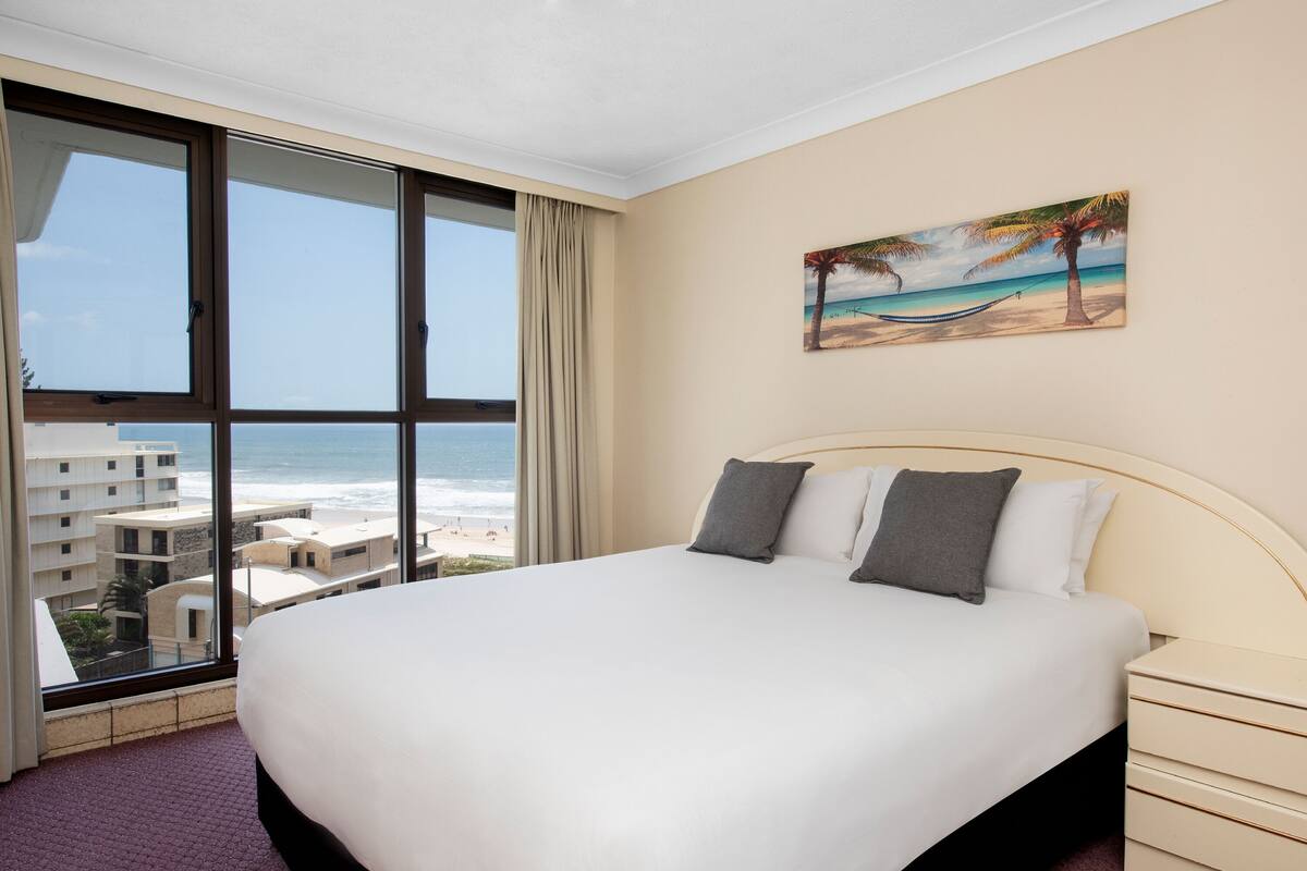 A hotel room with a large bed, a painting of a beach scene, and a view of the ocean through floor-to-ceiling windows.
