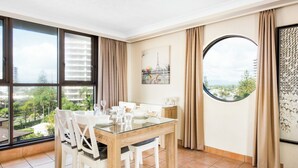 A dining area with a glass table, white chairs, and a view of greenery and buildings through large windows.