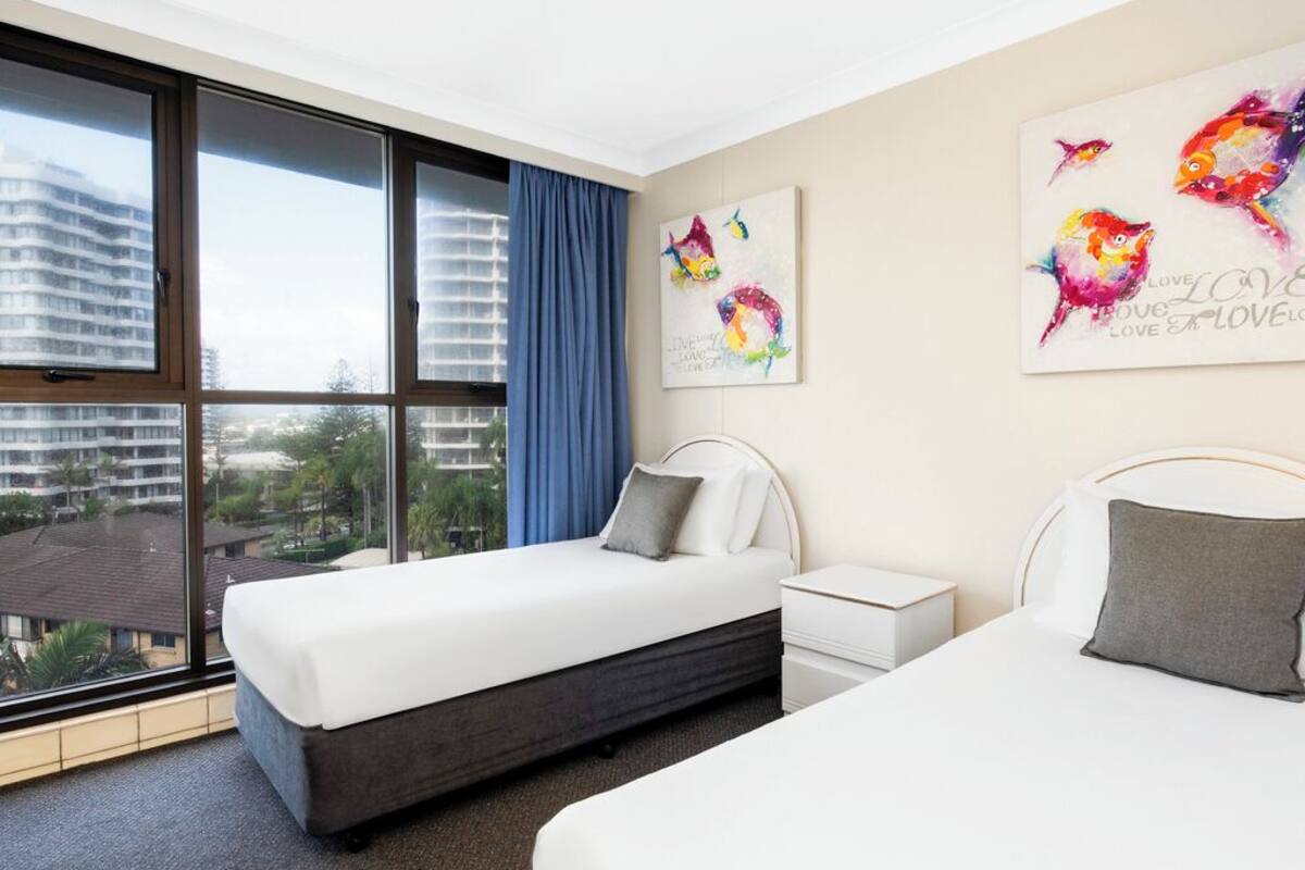 A hotel room with two beds, a large window with a view of buildings, and artwork on the wall.