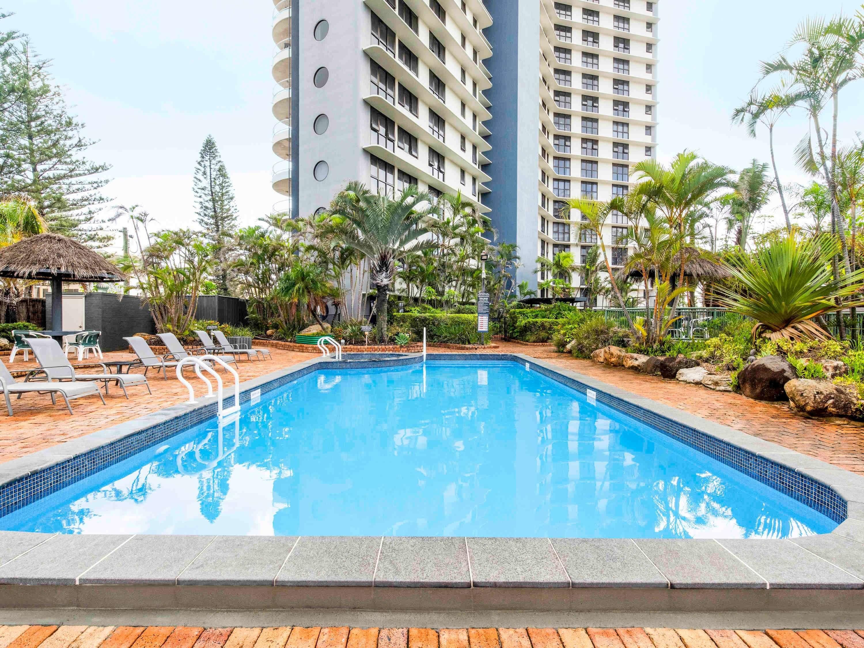 A swimming pool surrounded by lounge chairs, palm trees, and a tall building in the background.
