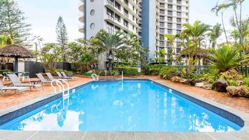 A swimming pool surrounded by lounge chairs, palm trees, and a tall building in the background.