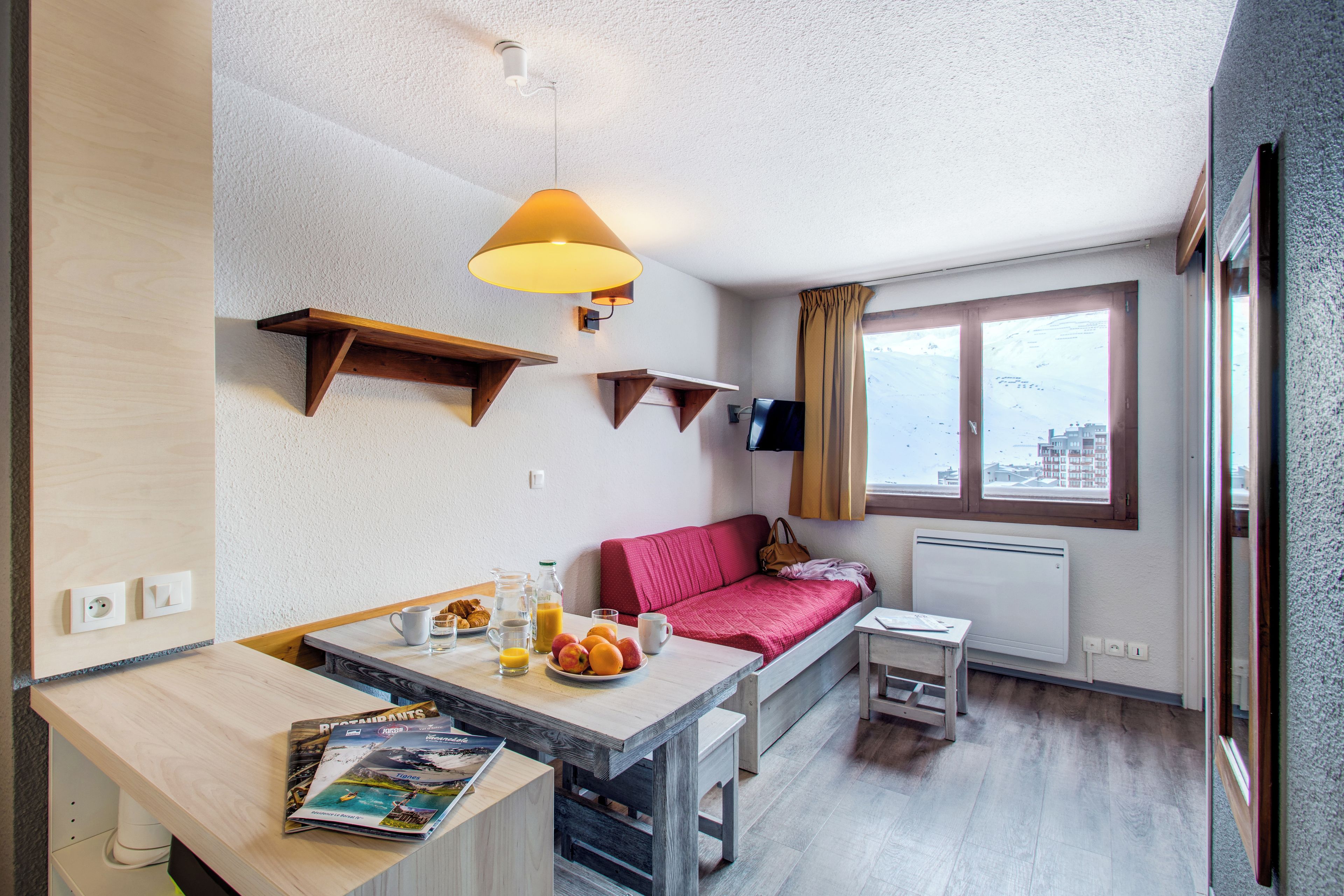 A compact living space with a kitchenette, a dining area, a red sofa, a small table, and a window with a city view.
