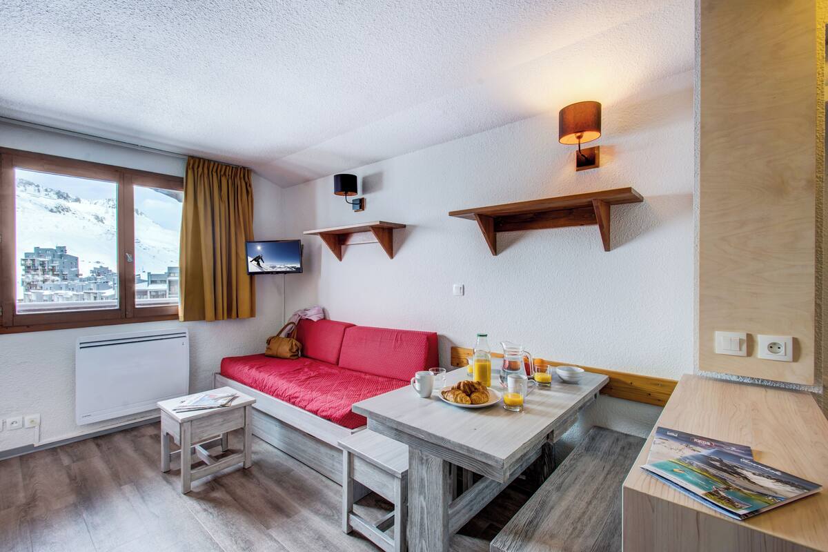 Una habitación moderna con un sofá rojo, una mesa de madera con alimentos para el desayuno, un televisor montado en la pared y vistas a la ciudad a través de amplias ventanas.
