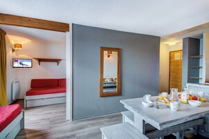 Una habitación moderna con pared gris, techo de madera, sofá rojo, mesa de comedor y alimentos para el desayuno, y un televisor en la pared.
