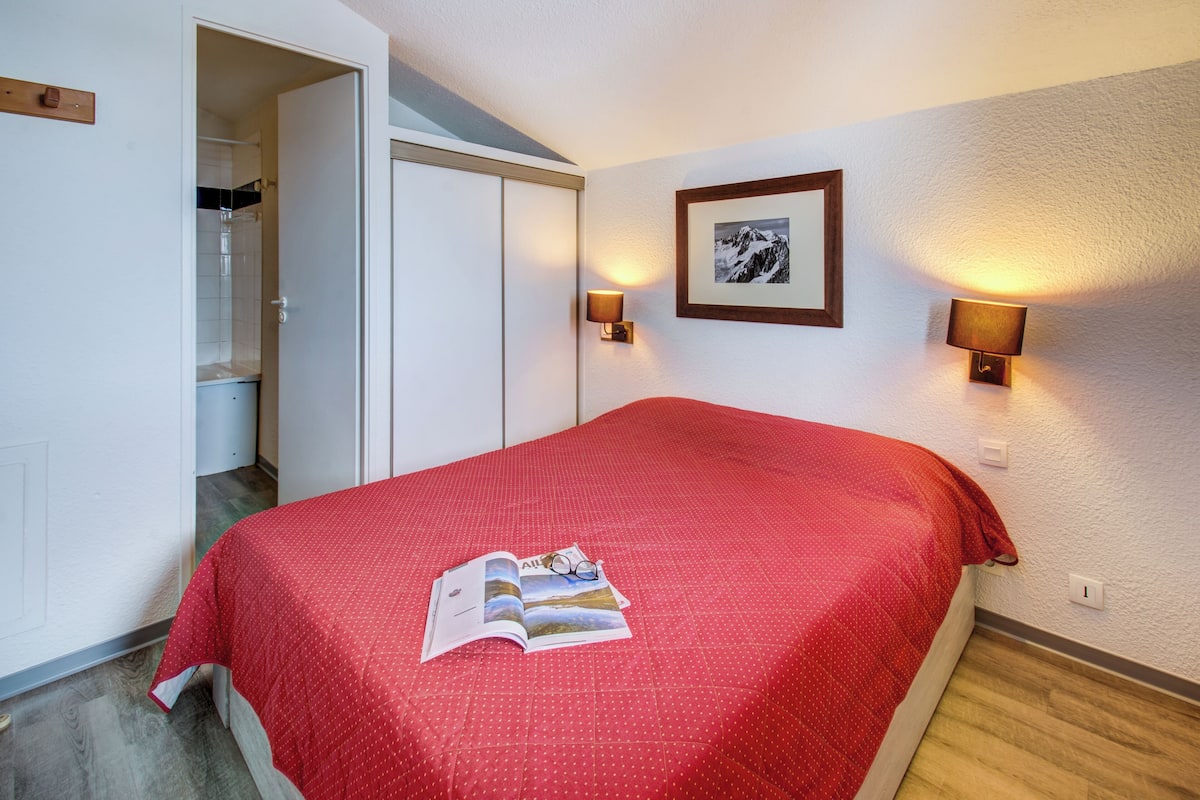 Un dormitorio con una colcha roja, un cuadro en la pared y dos lámparas fijadas a la pared.