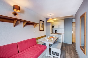 A compact living space with a red sofa, a dining table with breakfast items, a kitchen area, and a wooden ceiling beam.