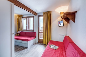 Una habitación con un sofá rojo, una ventana con cortinas amarillas, techo de madera y una lámpara fijada a la pared.
