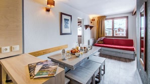 A modern living room with a wooden dining table, a red sofa, and a large window with a view of houses. - Résidence Le Borsat IV - Vacanceole (Tignes)