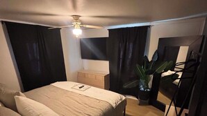 A compact hotel room with a bed, a desk, a chair, a potted plant, and a ceiling fan.