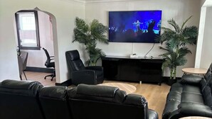 A modern living room with a large flat-screen TV, leather sofas, a wooden coffee table, and two potted plants.