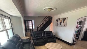 A living room with a black leather sofa, two wooden coffee tables, a staircase with string lights, and a view of the kitchen.