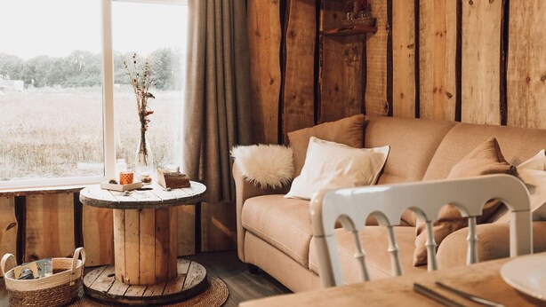 Ein gemütliches Zimmer mit Holzwand, einem runden Holztisch, einem beigen Sofa und einem Fenster mit Blick auf ein Feld.
