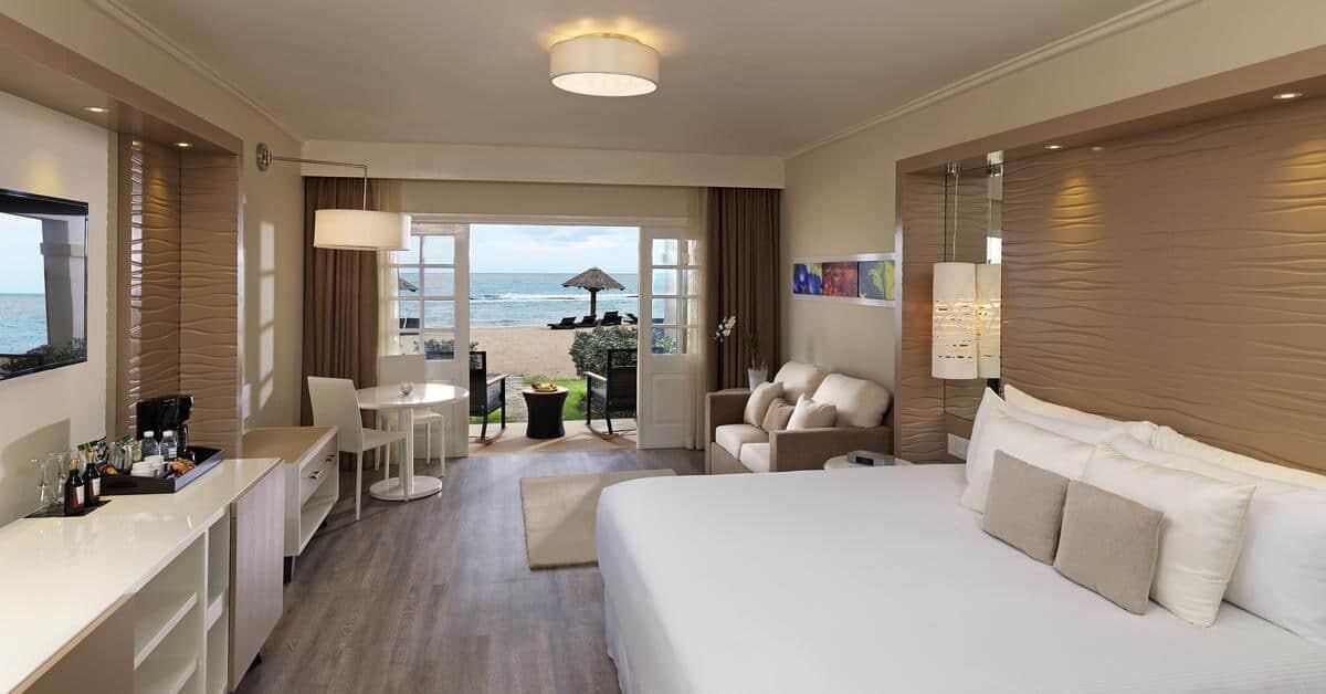 A hotel room with a bed, a desk, a chair, a small table, and a view of the beach.