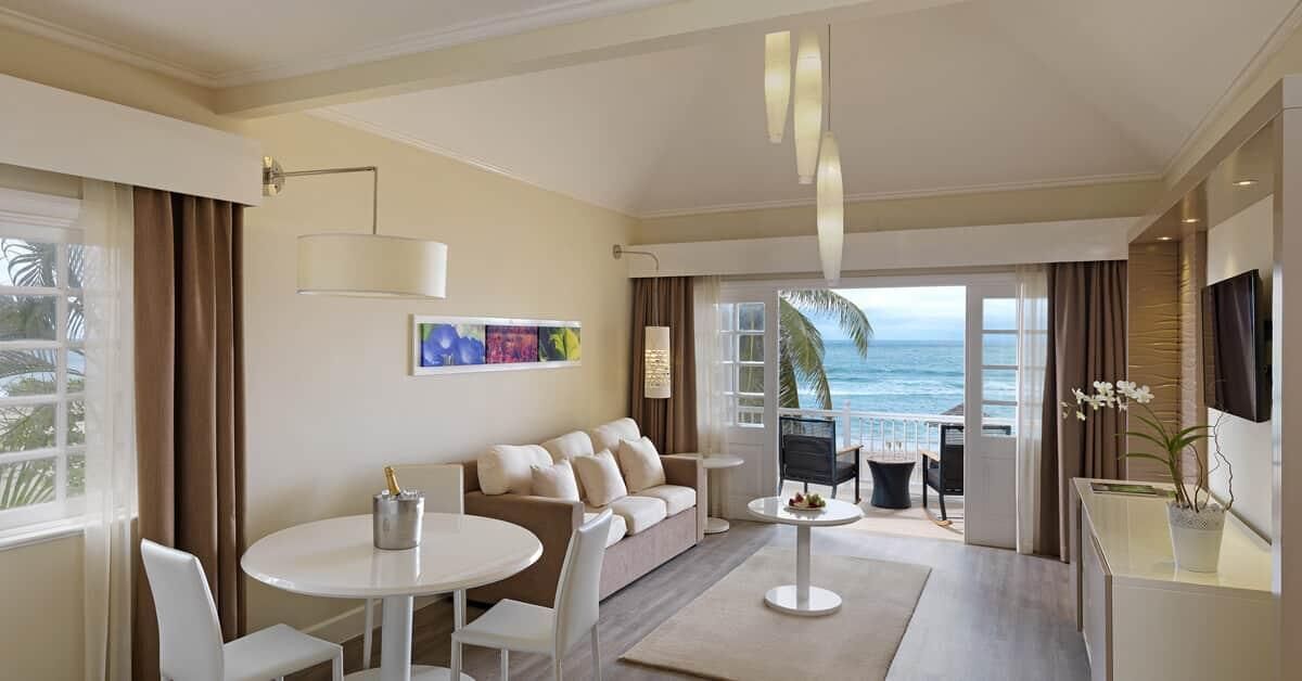 A modern hotel room with a balcony, a sofa, a dining table, and a TV.