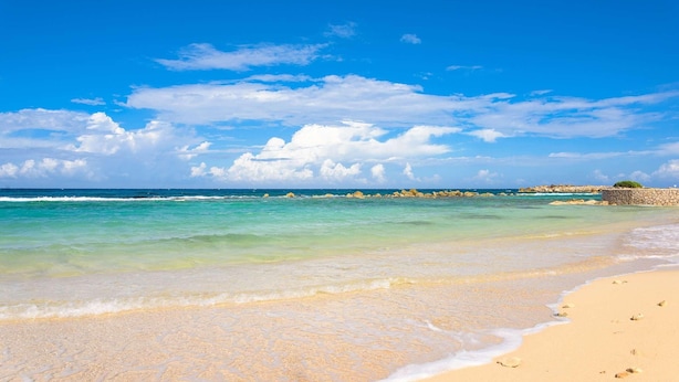 A beach with clear turquoise water, a sandy shore, and a distant rocky outcrop.