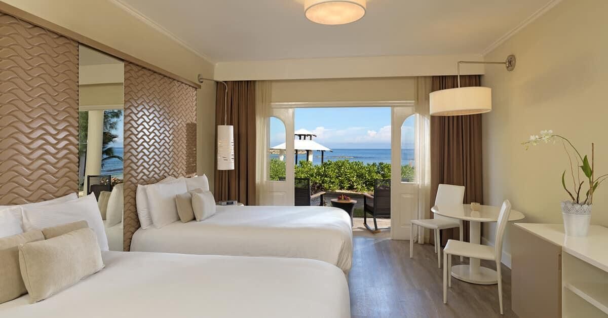 A hotel room with two beds, a desk, a chair, and a view of the ocean.