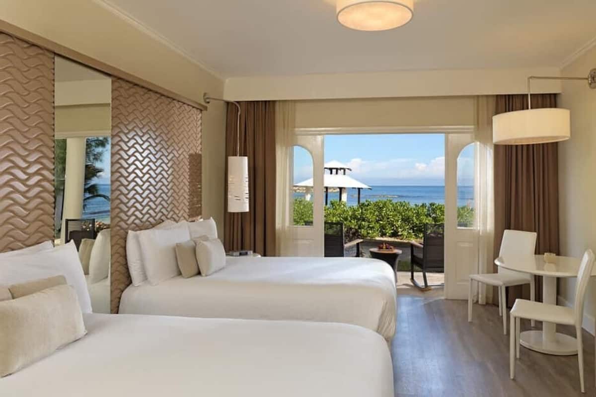 Quarto de hotel com duas camas, uma escrivaninha, uma cadeira e vista para o oceano.