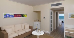 A living room with a beige sofa, a small round table with champagne glasses, a minibar, and a view of the ocean through an open door. - Melia Braco Village (Rio Bueno)