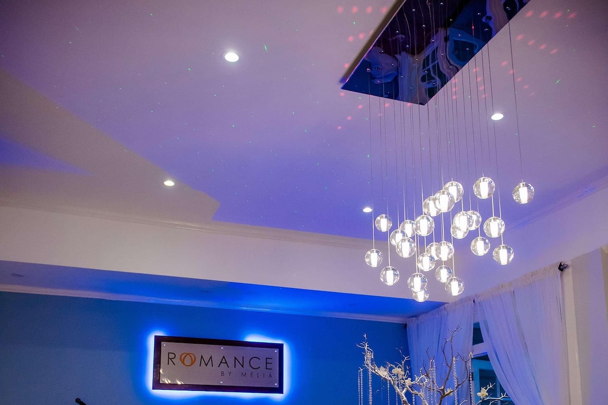Uma sala de estar moderna com um sofá, mesa de centro e objetos decorativos. Uma placa na parede diz 'ROMANCE BY MELIA'.
