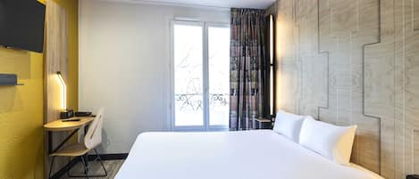 A hotel room with a large bed, a desk with a chair, a television mounted on the wall, and a window with curtains.