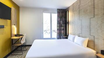 A hotel room with a large bed, a desk with a chair, a television mounted on the wall, and a window with curtains.