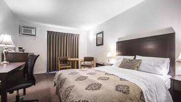 A hotel room with a bed, a desk, two chairs, a small table, and a nightstand with a lamp.