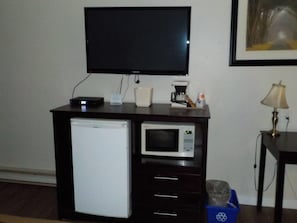 A room with a flat-screen TV mounted on the wall, a small refrigerator, a microwave, a coffee maker, and a trash bin.