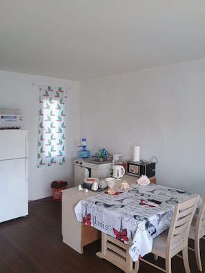 Uma pequena cozinha compacta com geladeira, micro-ondas e pia. Há uma mesa com toalha e cadeiras, e uma lixeira pequena.