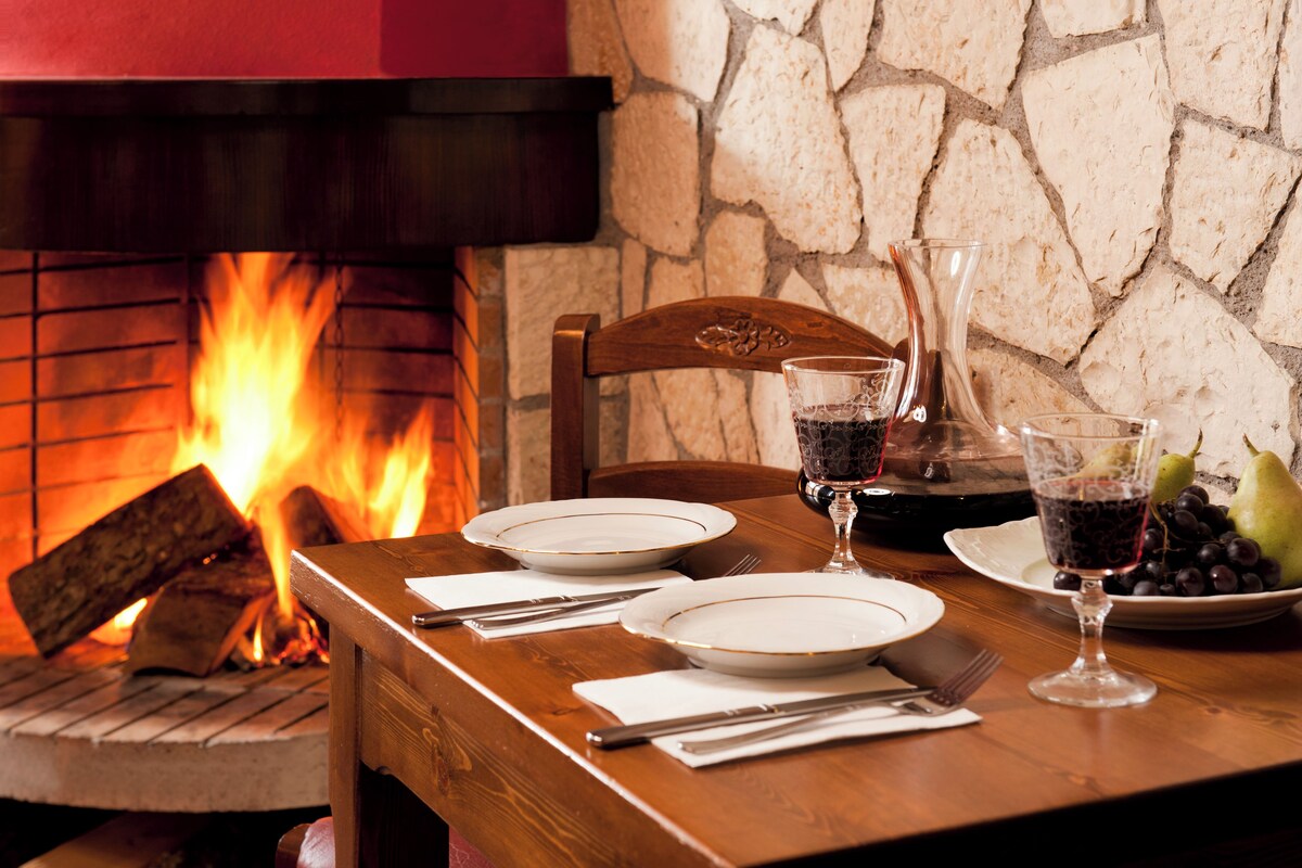 Una mesa de comedor dispuesta con platos, vasos, cubiertos y un frutero, con una pared de piedra y una chimenea al fondo.
