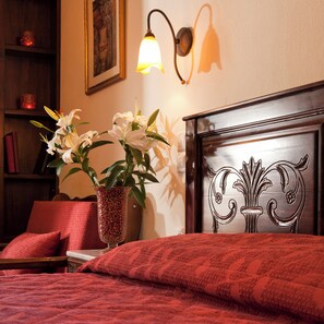 Una cama bien tendida con una colcha roja estampada, un jarrón con flores blancas, una lámpara de pared y un cuadro en la pared.