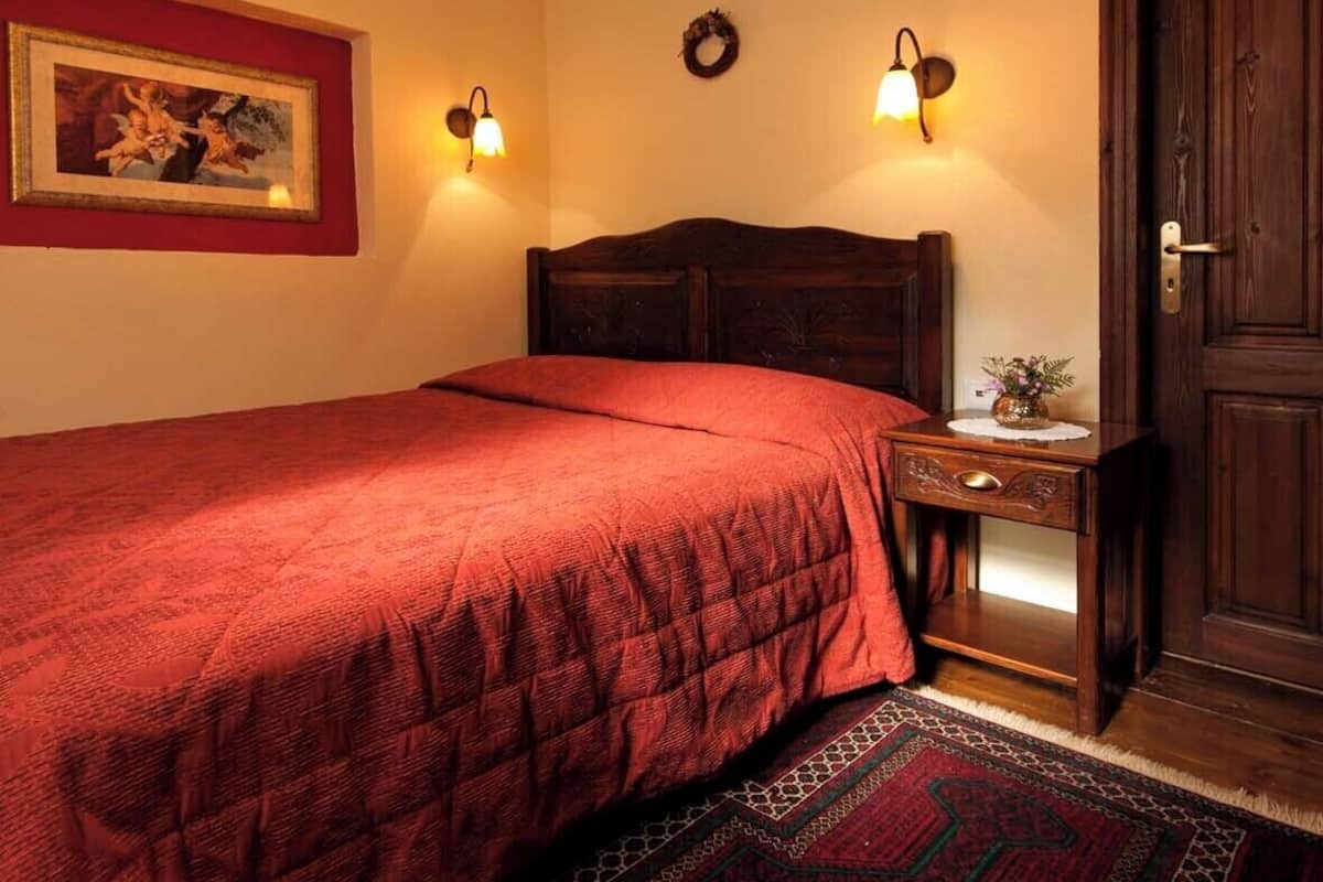 Ein Schlafzimmer mit einem großen Bett, einem hölzernen Kopfteil, einer roten Bettdecke, einem gemusterten Teppich, einem kleinen Tischchen mit einer Vase und zwei Wandleuchten.