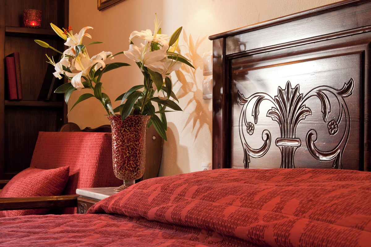 A neatly made bed with a red patterned bedspread, a vase with white flowers, a wall-mounted light fixture, and a framed picture on the wall.