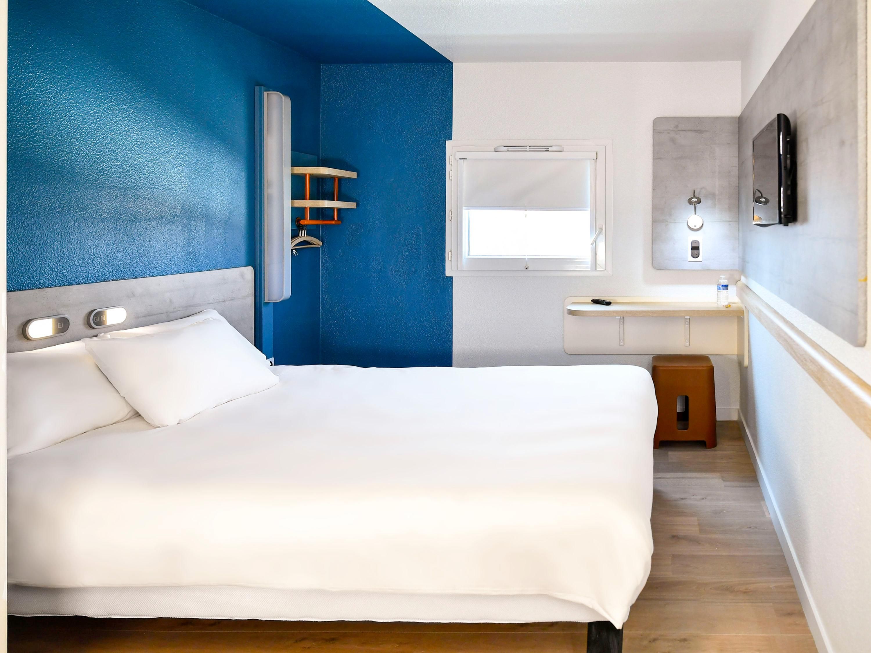 Quarto de hotel com cama, pequena escrivaninha, janela e área de chuveiro.