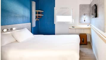 Quarto de hotel com cama, pequena escrivaninha, janela e área de chuveiro.