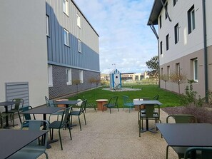 An outdoor patio area with tables and chairs, a small garden, and a building with a modern design. - Ibis Budget Le Treport Mers les Bains (Mers-les-Bains)
