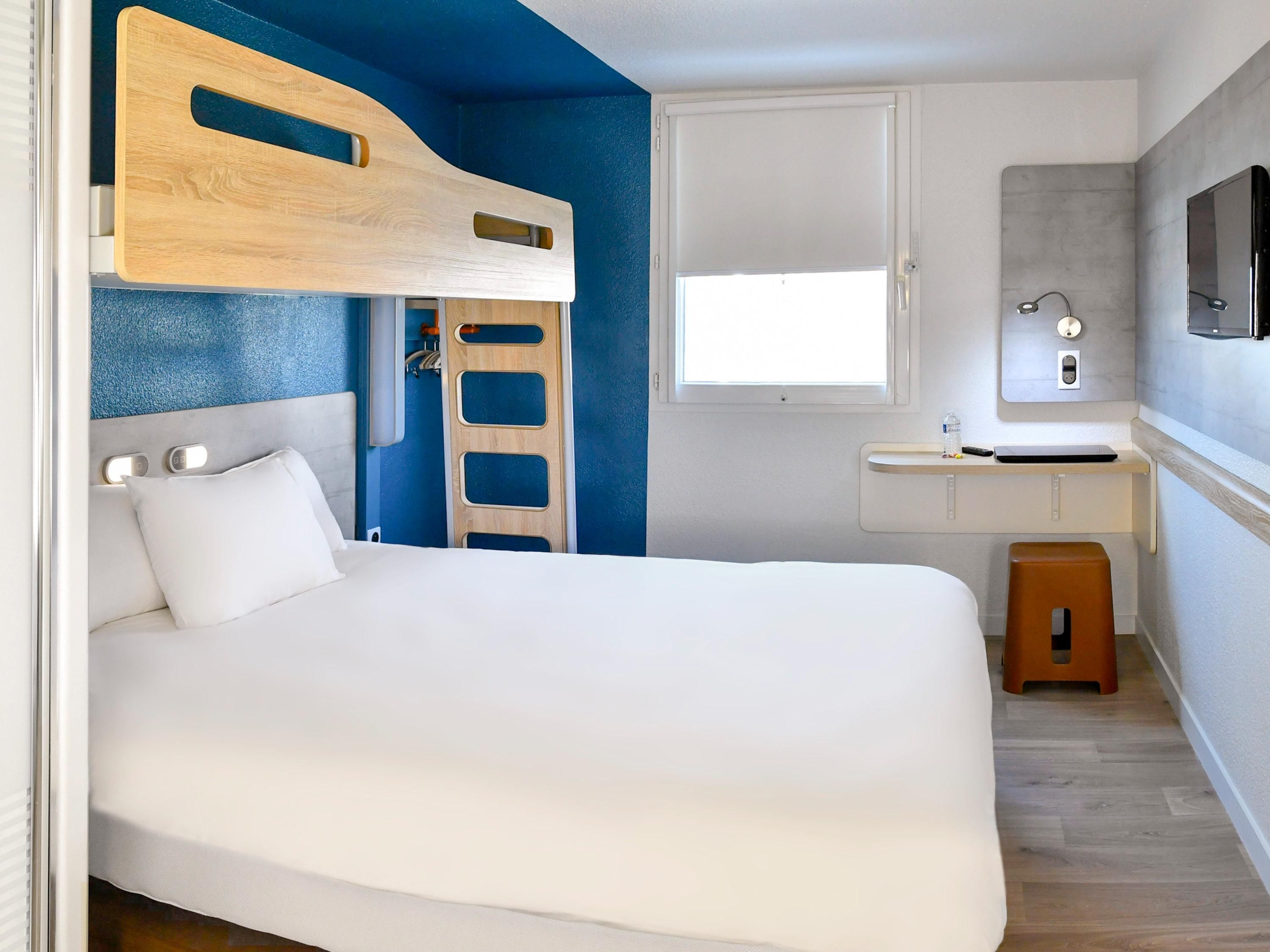 Una cama tipo litera con cabecera de madera, una mesita de noche blanca, un taburete pequeño, una ventana y un baño con espejo y lavamanos.