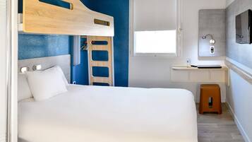 Una cama tipo litera con cabecera de madera, una mesita de noche blanca, un taburete pequeño, una ventana y un baño con espejo y lavamanos.