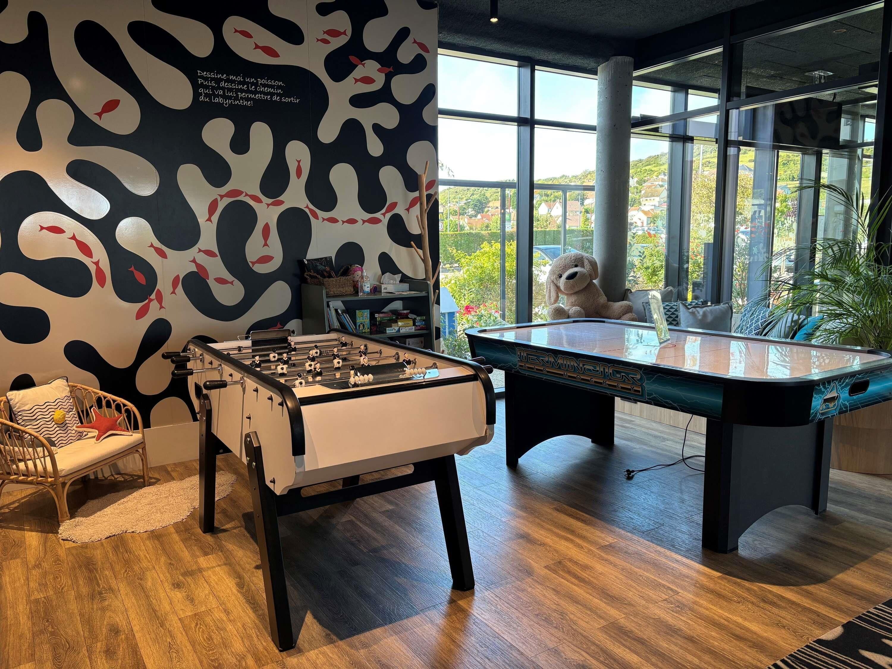 A room with a foosball table, a pool table, a wicker chair, and a large window with a view of greenery.