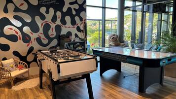 A room with a foosball table, a pool table, a wicker chair, and a large window with a view of greenery.