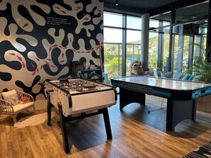 A room with a foosball table, a pool table, a wicker chair, and a large window with a view of greenery. - Ibis Budget Le Treport Mers les Bains (Mers-les-Bains)