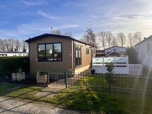 Ein kleines, eingeschossiges Gebäude mit Holzwänden und großen Fenstern, umgeben von einem eingezäunten Garten und weiteren Wohnhäusern.