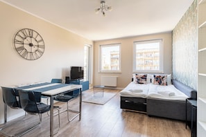 Ein modernes Zimmer mit Esstisch, Stühlen, Schlafsofa, Fernseher und großen Fenstern.