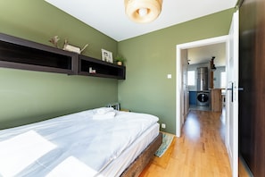 Un dormitorio con cama, piso de madera, pared verde y un pasillo que lleva a una cocina.